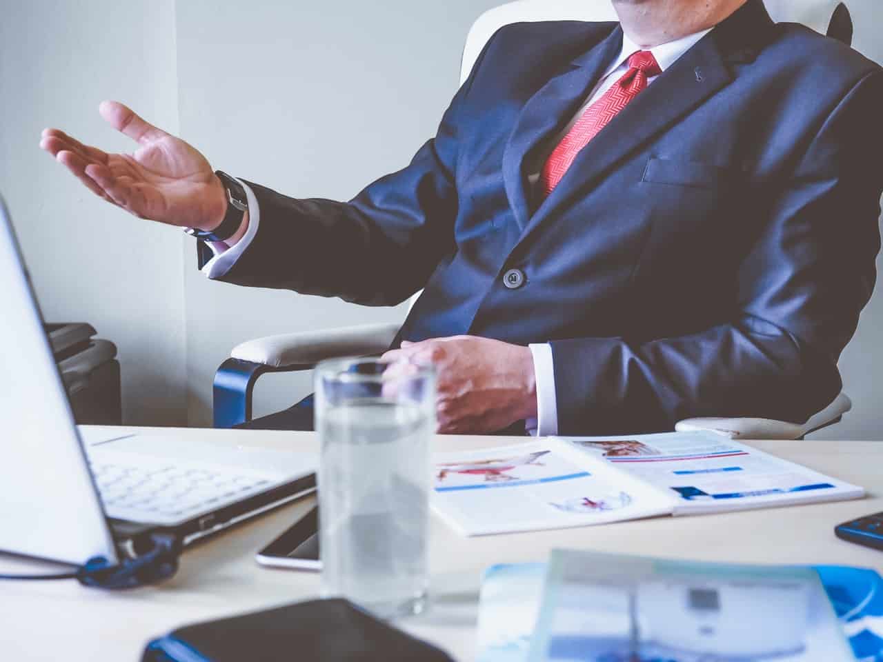 A man wearing a black suit gesturing to a laptop